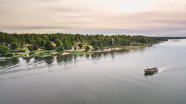 Flygbild över Kristinehamns skärgård under en solnedgång. En ensam båt glider fram över spegelblankt vatten. Längst bort i bort i bild syns strandkanten, mede båtbryggor och den angränsande skogen.